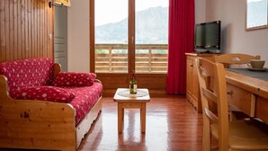 A cozy room with a wooden sofa, a small table with a bottle, a TV on a wooden stand, and a view of mountains through a large window. - Cozy 2-room apartment for 4, mountain view balcony, pets allowed. (Le Dévoluy)