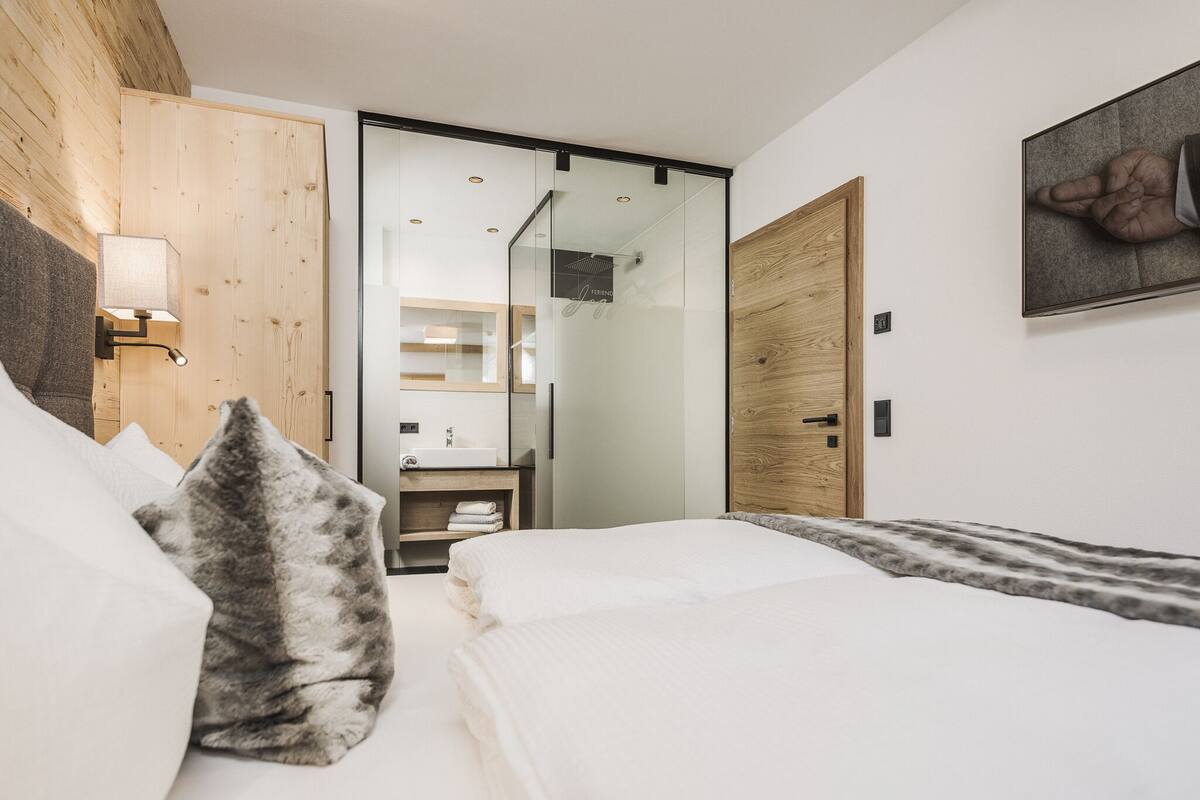 A modern hotel room with a wooden headboard, a large bed with white linens, a bedside table with a lamp, and a view into the bathroom.