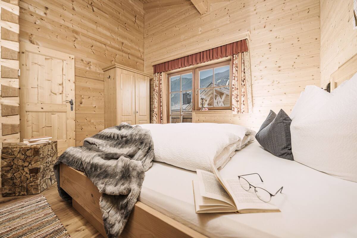 A cozy wooden cabin bedroom with a bed, a window with curtains, and a small wooden chest.