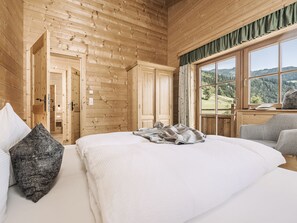 A wooden cabin bedroom with a bed, pillows, a window with a view of the countryside, and a wooden wardrobe.