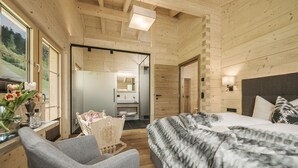 Une chambre aménagée dans une cabane en bois, avec un lit, une chaise, un bureau agrémenté d’un vase de fleurs et une salle de bain visible à travers une porte ouverte.