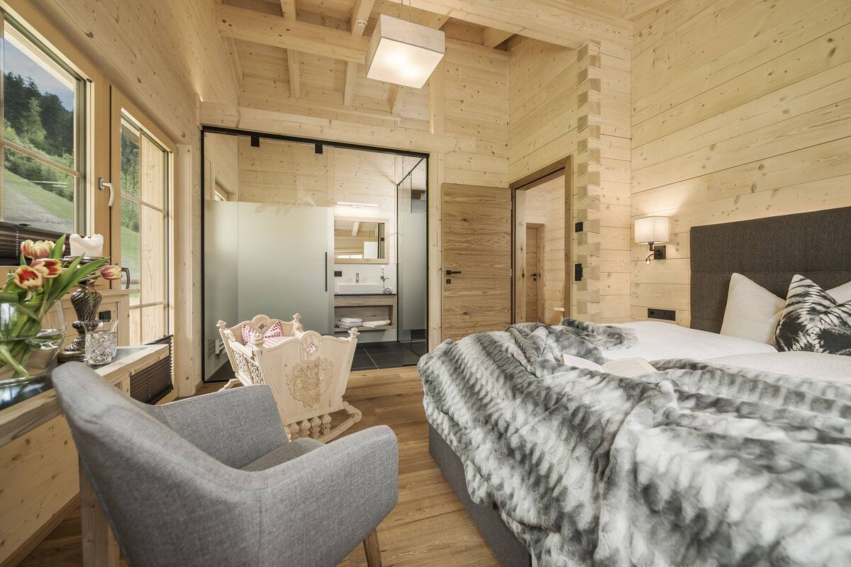 A wooden cabin bedroom with a bed, a chair, a desk with a vase of flowers, and a bathroom visible through an open door.