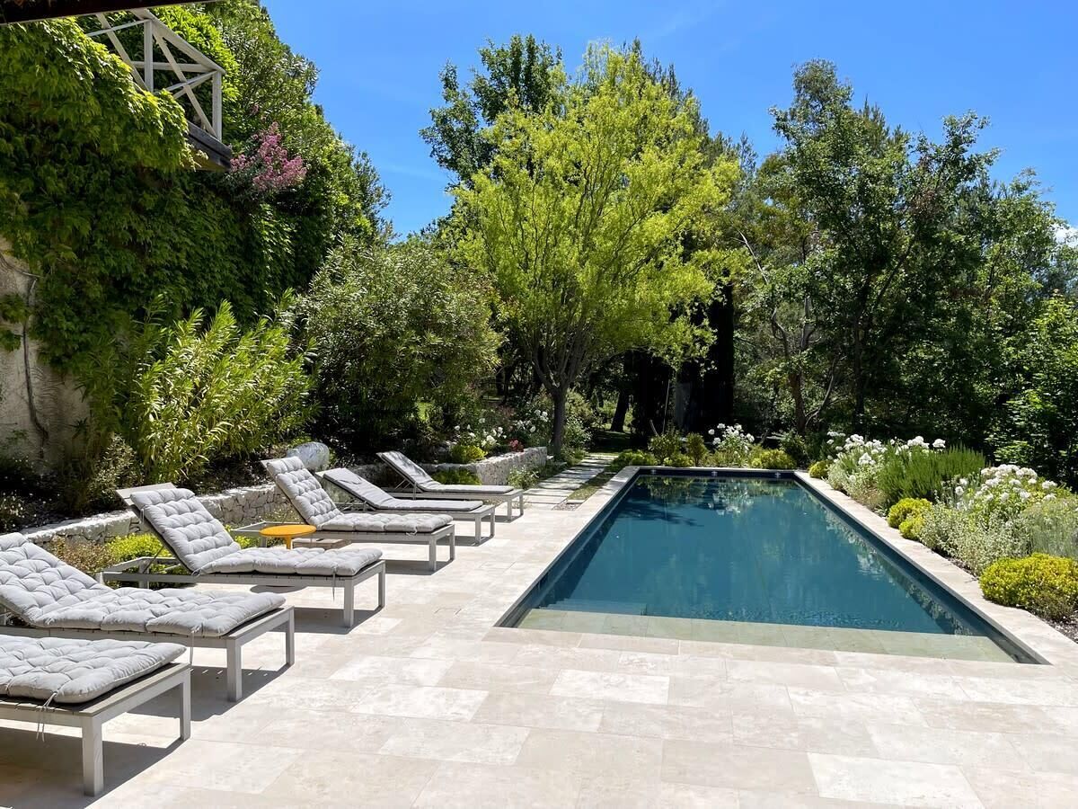 Une piscine rectangulaire, bordée d’une terrasse carrelée, entourée de chaises longues et de verdure.