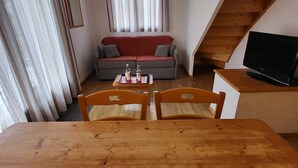 A cozy living room with a wooden dining table, two chairs, a red sofa, a TV on a wooden stand, and a staircase.
