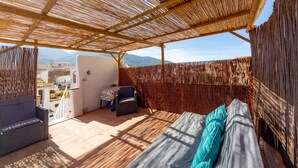 A covered outdoor seating area with a sofa and chairs, a table, and a view of distant buildings and mountains. - Traditional Spanish casa in the heart of town. (Órgiva)