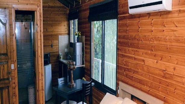 A wooden cabin room with a bed, a small table with chairs, a kitchen area, and an air conditioning unit. - Pearl Of The North (Hazor Haglilit)