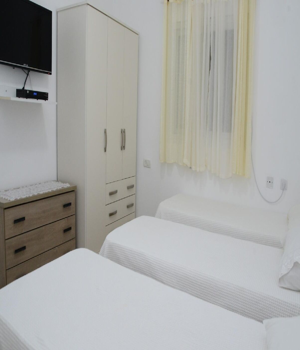Un dormitorio con dos camas individuales, un armario, un televisor montado en la pared y un enchufe.