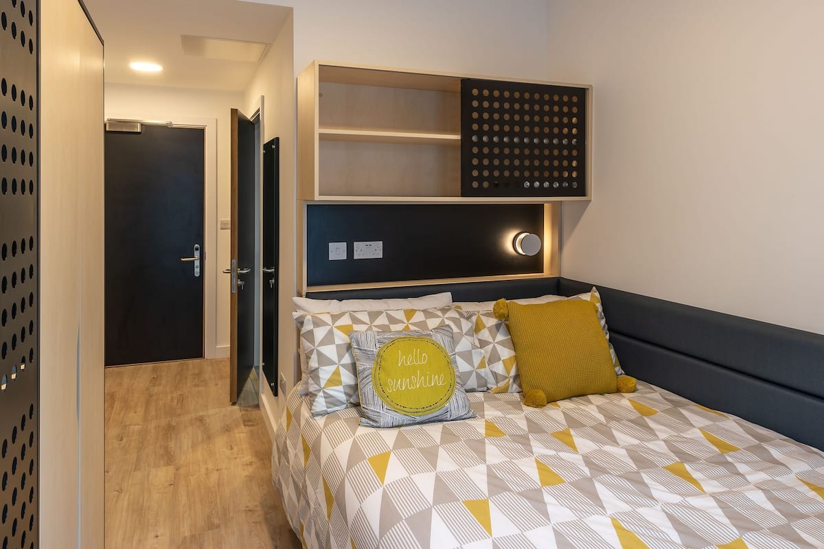 A modern bedroom with a geometric-patterned bedspread, a built-in shelf with a decorative panel, and a visible door leading to another room.