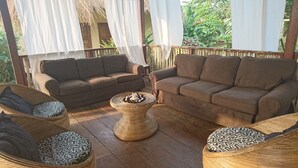 A covered outdoor seating area with two sofas and two wicker chairs, a woven coffee table, and white curtains. - Eden Lodge Kribi (Kribi)