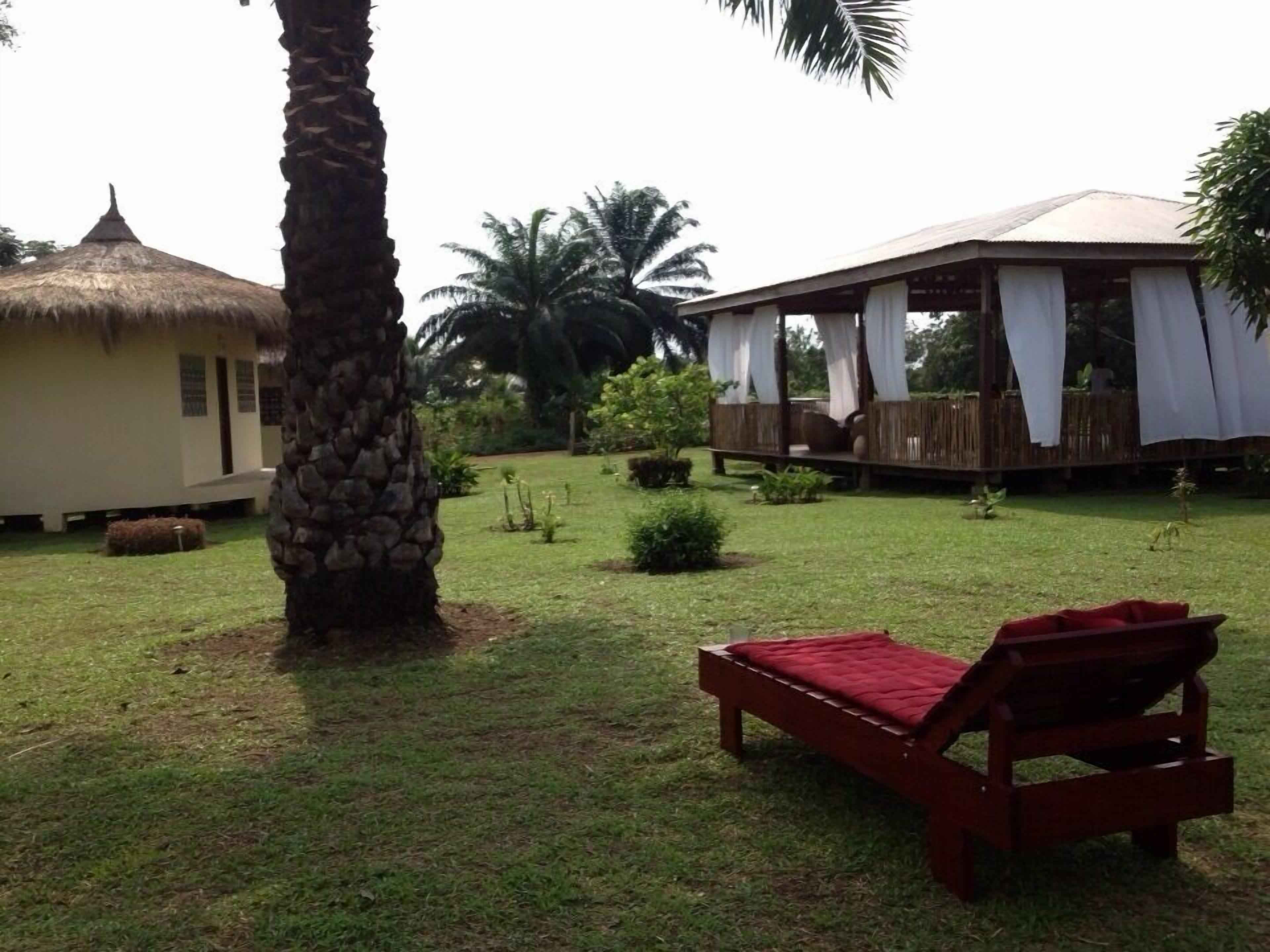 A grassy area with a thatched-roof structure, a wooden deck with white curtains, and a red lounge chair.