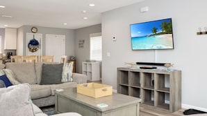 A living room with a beige sofa, a coffee table, and a TV on the wall. - Steps To The Beach, Near Killer Seafood ~ Oleander Place (Mexico Beach)
