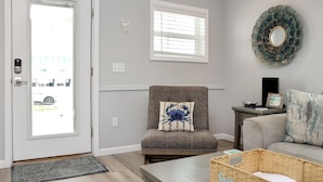 A living room with a gray sofa, a chair with a crab pillow, a wooden coffee table, a wicker basket, a wall-mounted mirror, and a window with blinds. - Steps To The Beach, Near Killer Seafood ~ Oleander Place (Mexico Beach)
