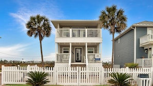 A two-story house with a balcony, surrounded by a white picket fence, palm trees, and a clear blue sky. - Steps To The Beach, Near Killer Seafood ~ Oleander Place (Mexico Beach)