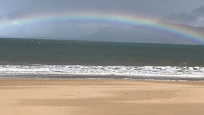A beach with a rainbow over the ocean and mountains in the background.