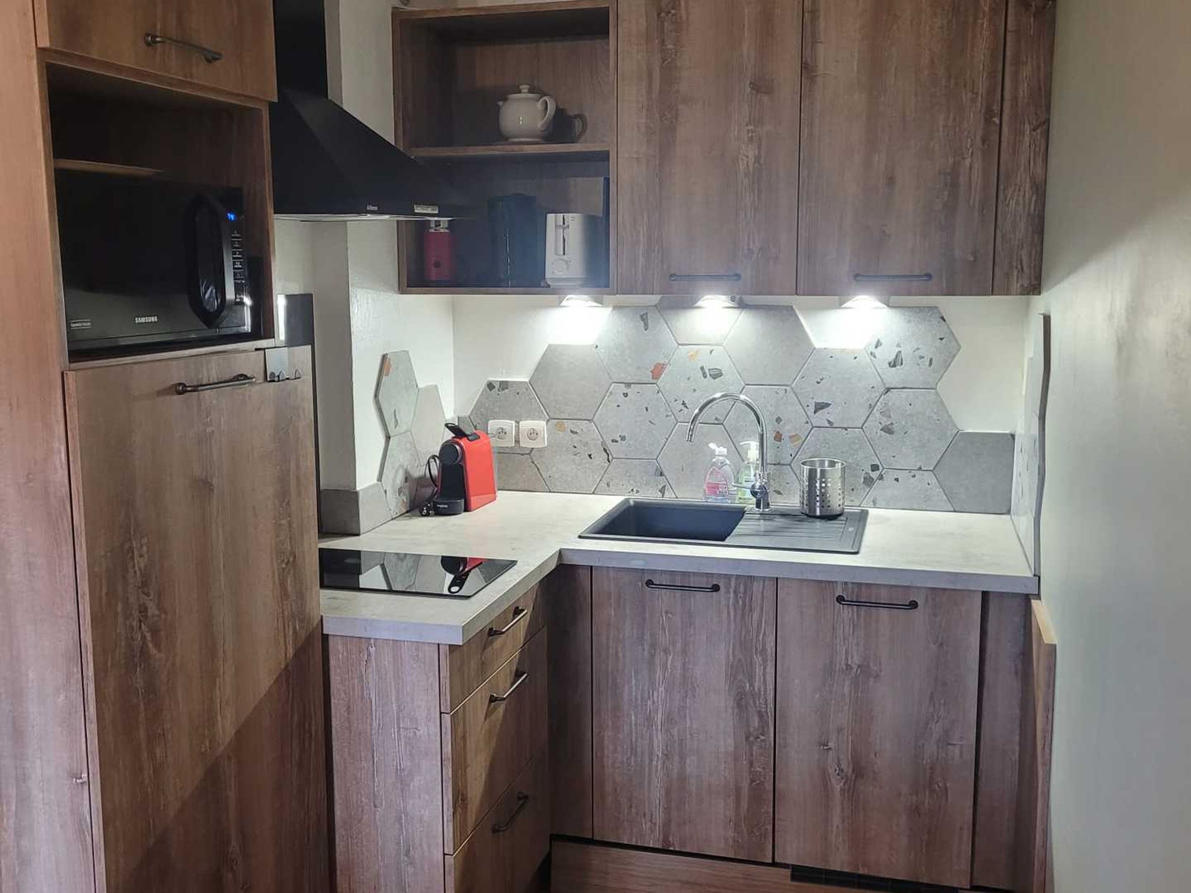 Een kleine keuken met houten kastjes, een magnetron, een waterkoker en een moderne betegelde achterwand.