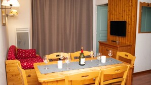 A cozy room with a wooden dining table set for four, a sofa with red pillows, a television on a wooden cabinet, and a large window with curtains. - 3-Room Apartment, 6 People, Pets Allowed, Balcony (Le Dévoluy)