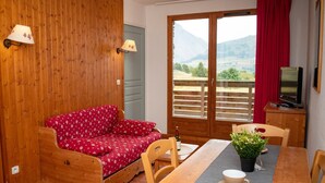 Een knus ingerichte kamer met een houten vloer, een rode bloemenbank, een houten eettafel met stoelen, een kleine potplant en een televisie op een houten kast.