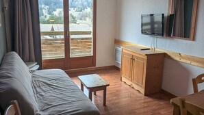 A room with a bed, a small table, a wooden cabinet, a TV, and a window with a view of a forest and mountains.