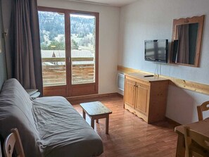A room with a bed, a small table, a wooden cabinet, a TV, and a window with a view of a forest and mountains. - 3-Room Apartment, 6 People, Pets Allowed, Parking (Le Dévoluy)