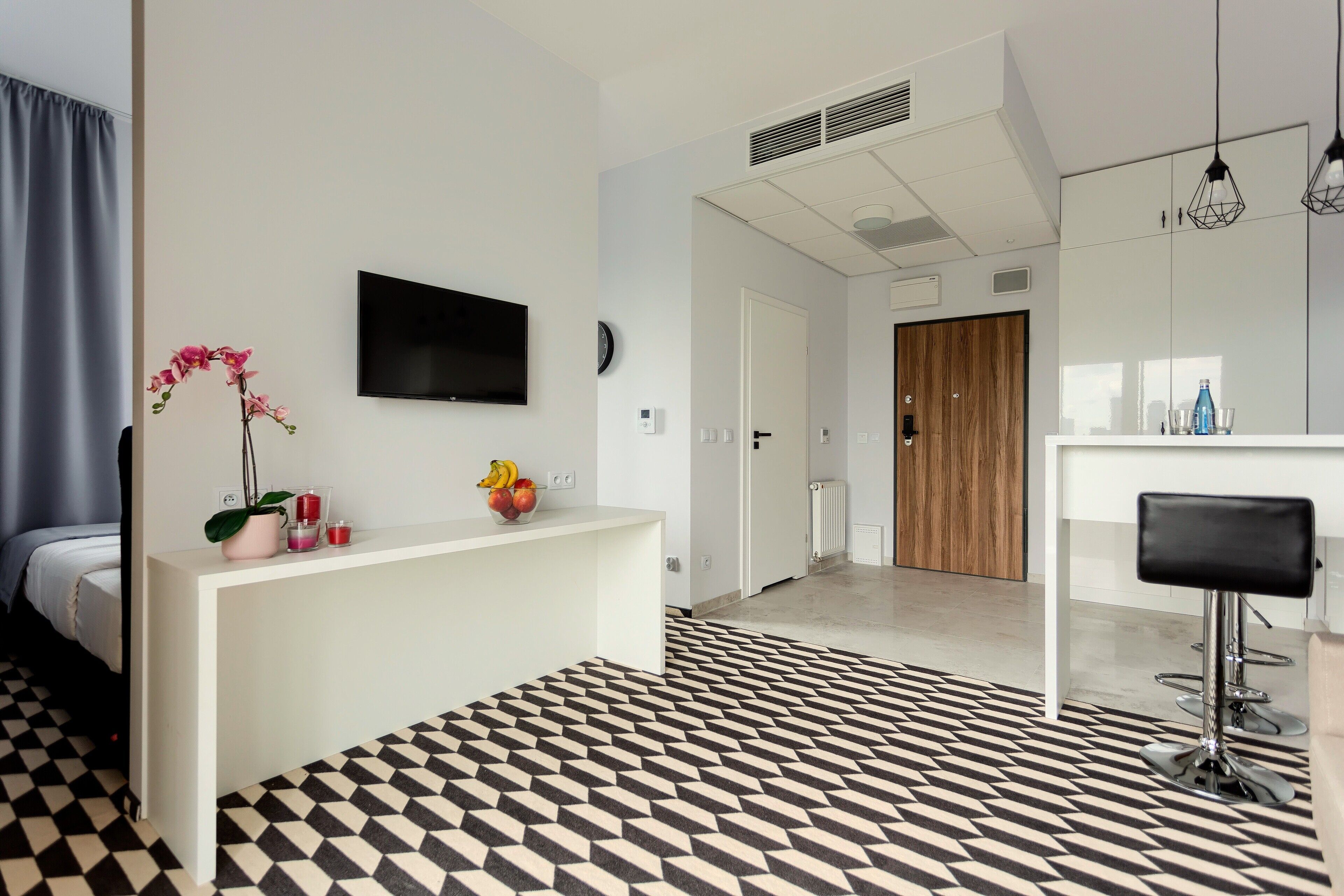 A modern hotel room with a geometric patterned floor, a white console table with decorative items, a flat-screen TV, a wooden door, and a bar area with a black bar stool.