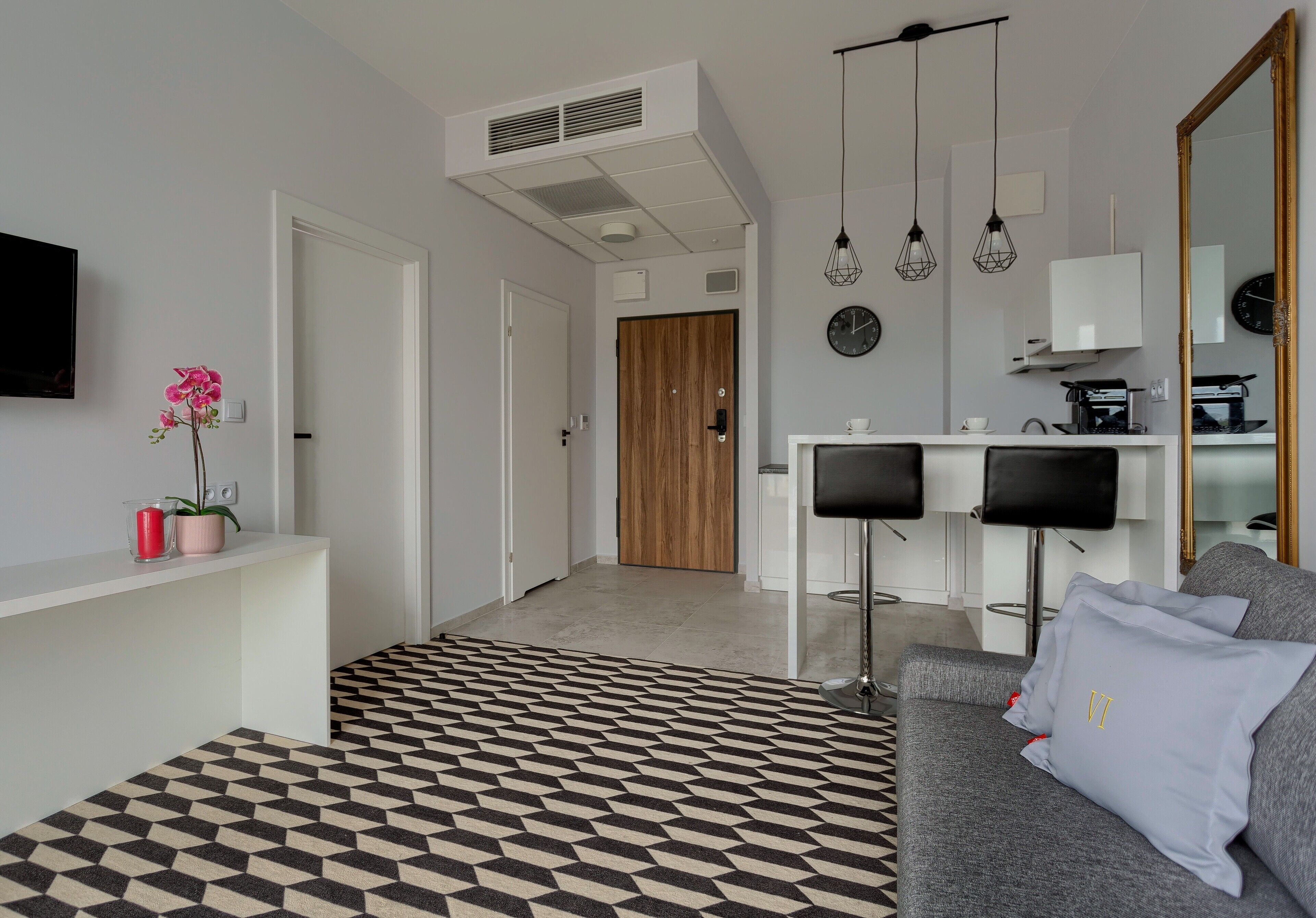 A modern living room with a geometric patterned rug, a grey sofa with a light grey pillow, a white console table with a pink vase and a red candle, and a wooden door in the background.