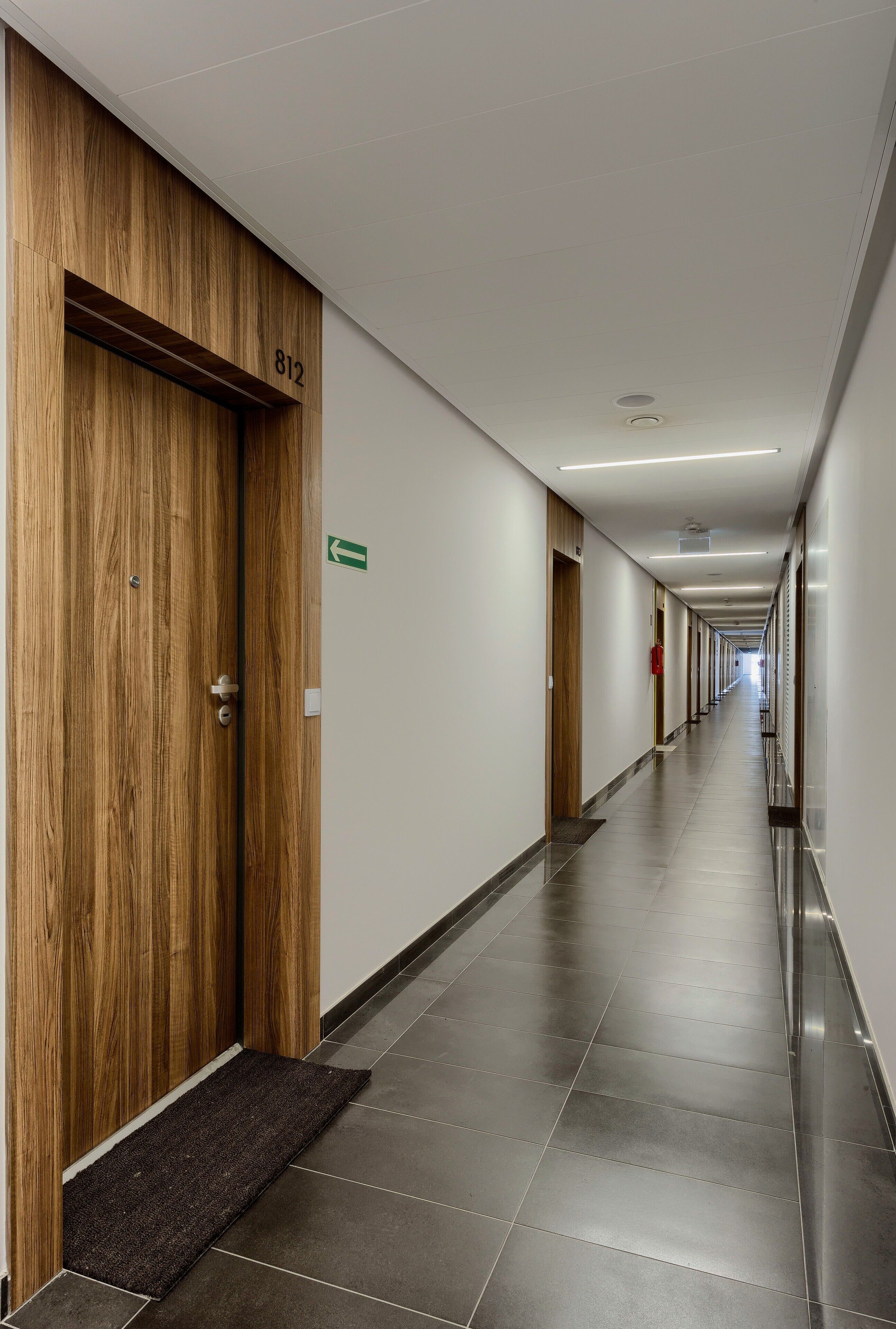 A long, narrow hotel corridor with wooden doors, numbered 812, and a green directional sign on the wall.
