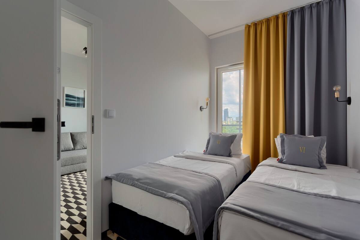 A bedroom with two beds, a window with curtains, a door leading to another room, and a patterned floor.