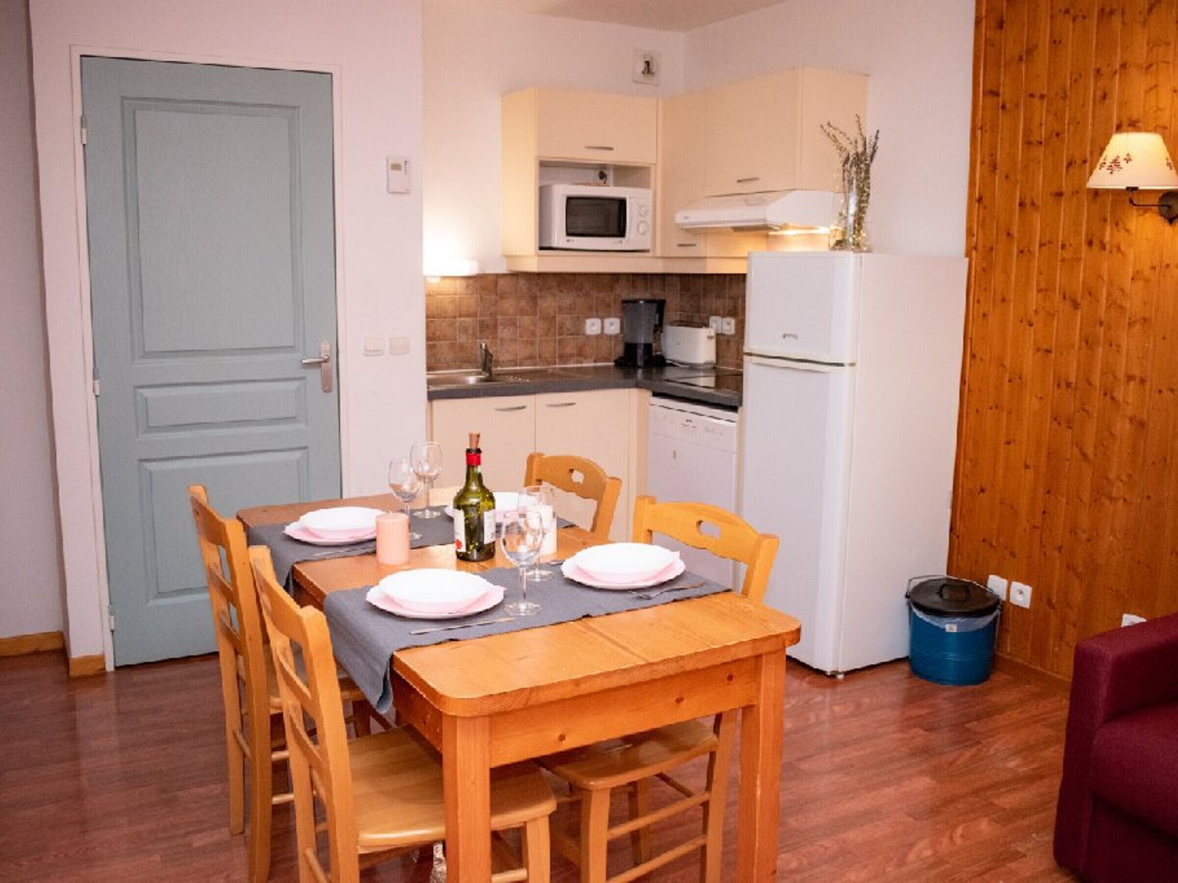 A compact kitchen with a dining table set for two, a microwave, a refrigerator, and a trash bin.