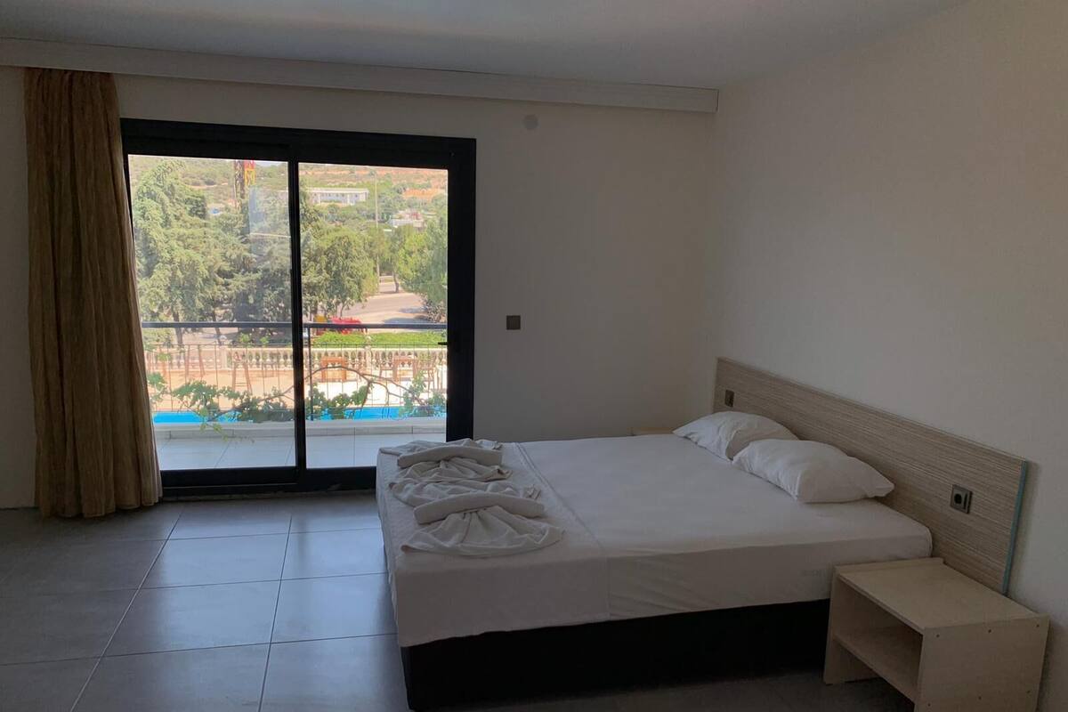 A hotel room with a bed, a small bedside table, and a view of a swimming pool and greenery outside.