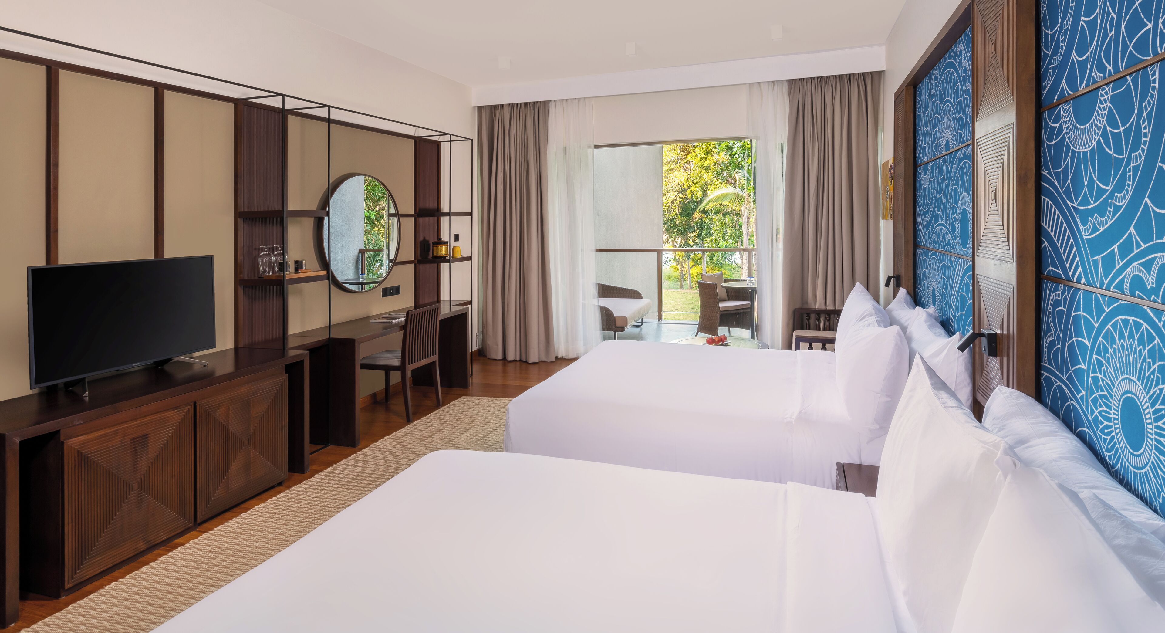 A hotel room with two beds, a TV, a desk, and a large window with a view of greenery.