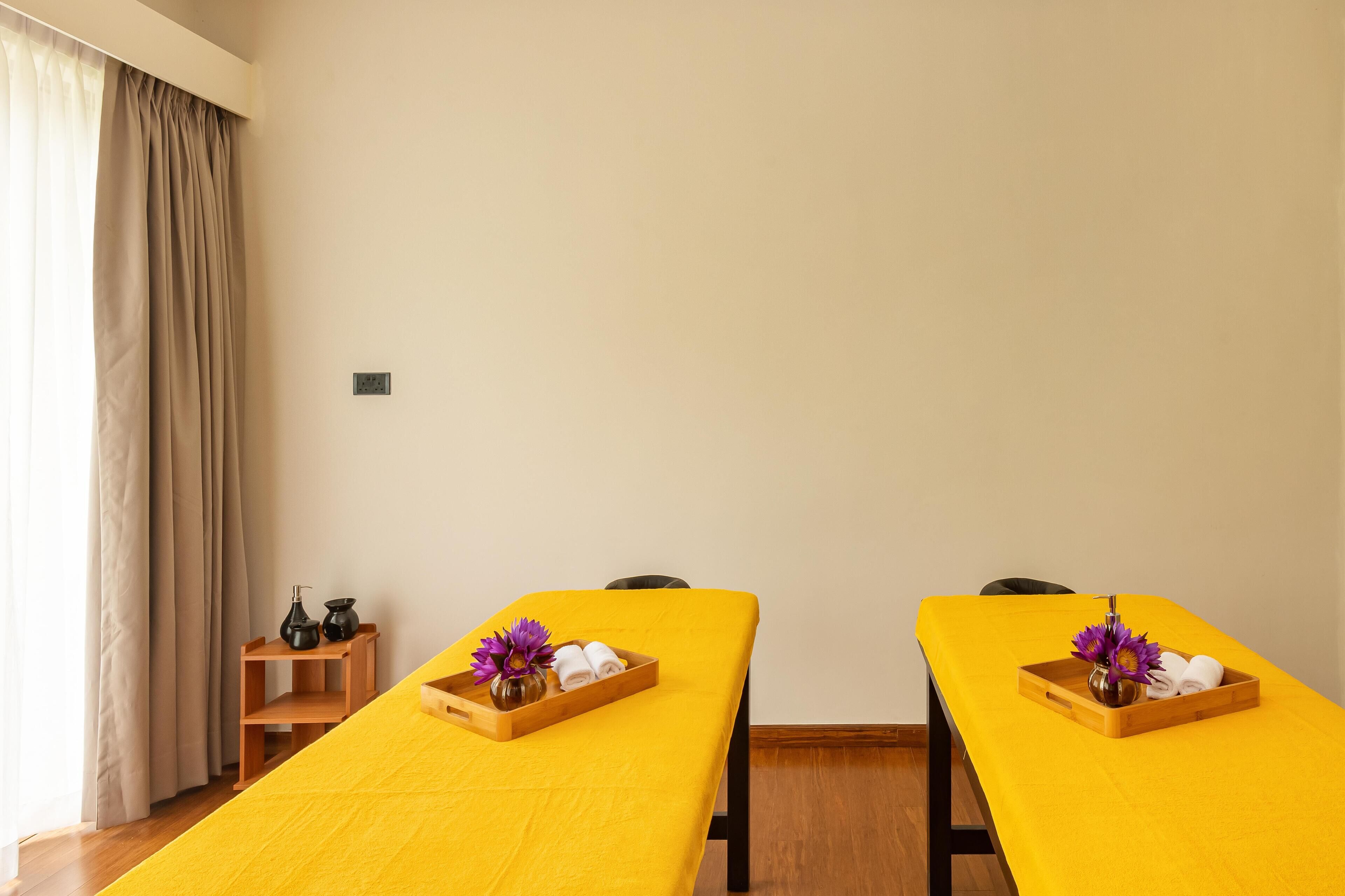 A room with two massage tables covered in yellow sheets, each with a tray of towels and a vase of purple flowers.