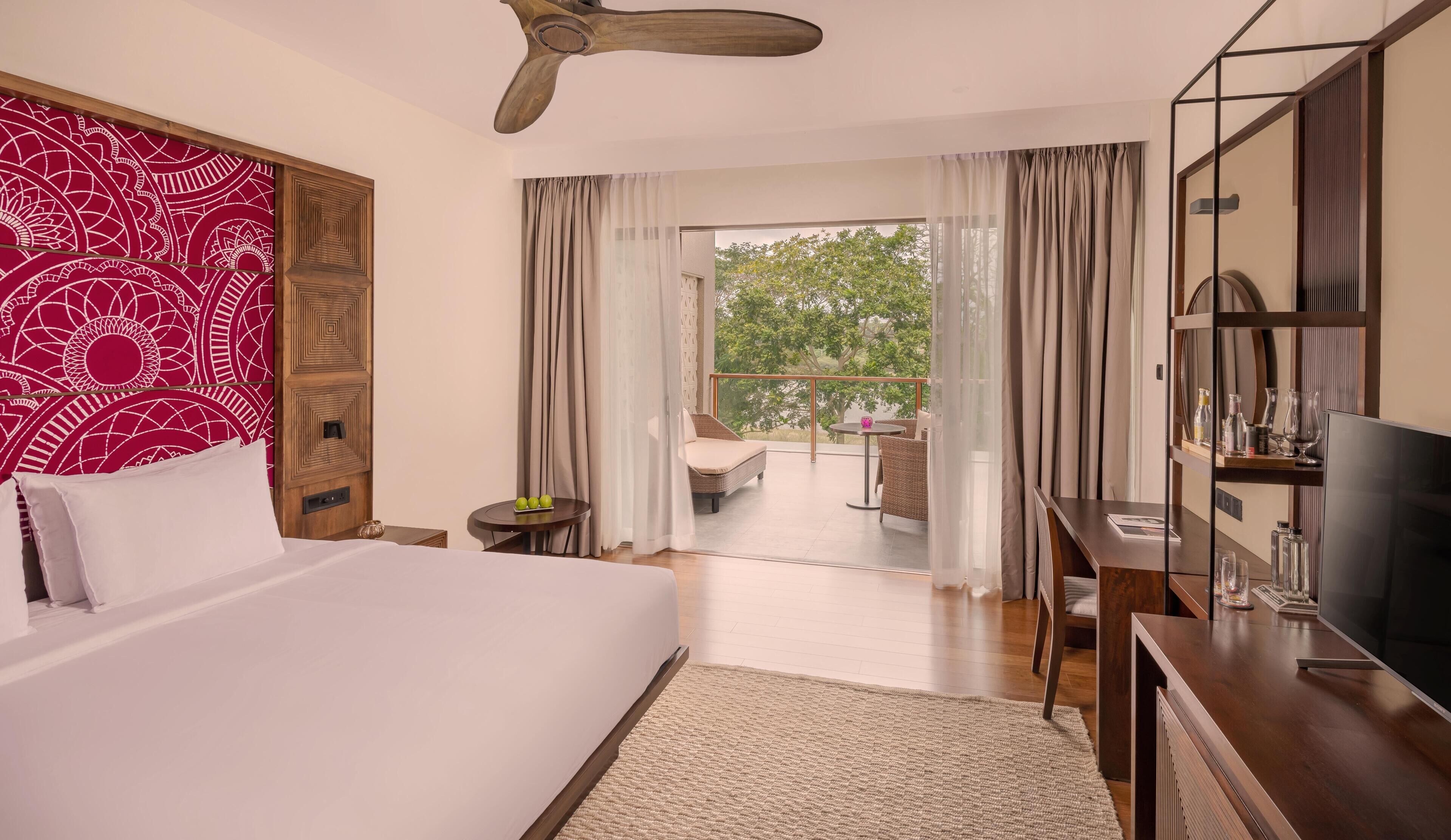 A hotel room with a large bed, a TV, a desk with a computer, a small table with apples, and a balcony with a chair and table.