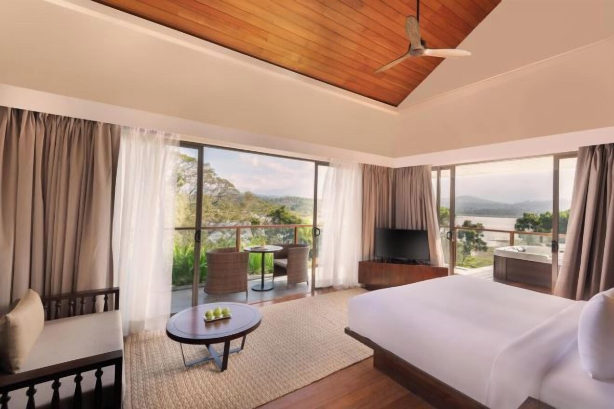 A hotel room with a large bed, a TV, a balcony with a table and chairs, and a view of the mountains.