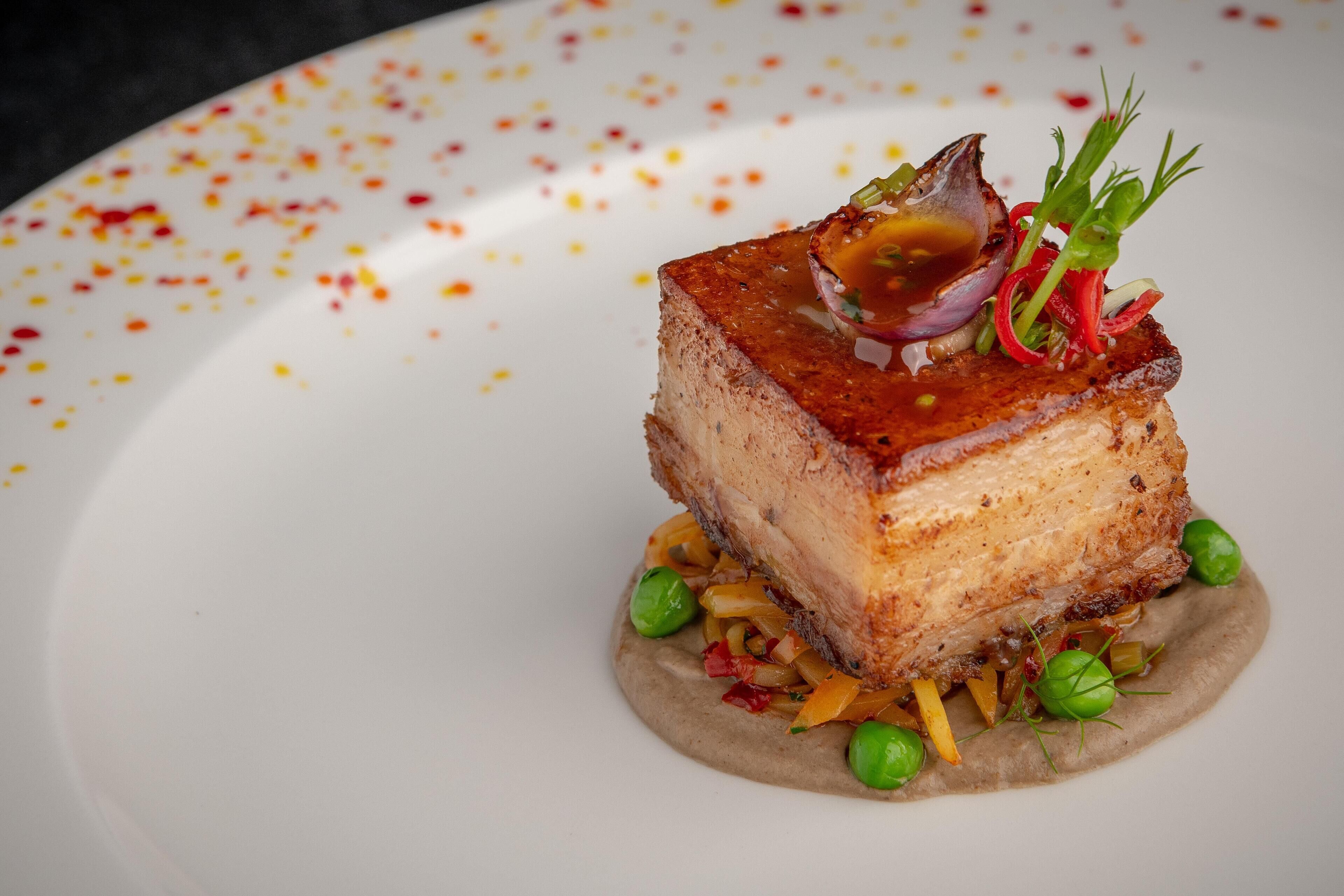 En fyrkantig rätt med en krispig yta, garnerad med gröna örter och en sås, serverad på en vit tallrik med ett prickigt mönster.
