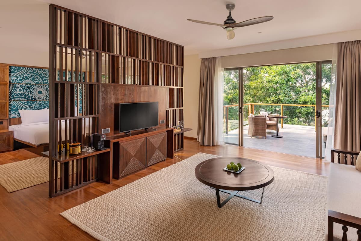 A modern living room with a large flat-screen TV, wooden decor, a round coffee table, and a balcony with outdoor furniture.