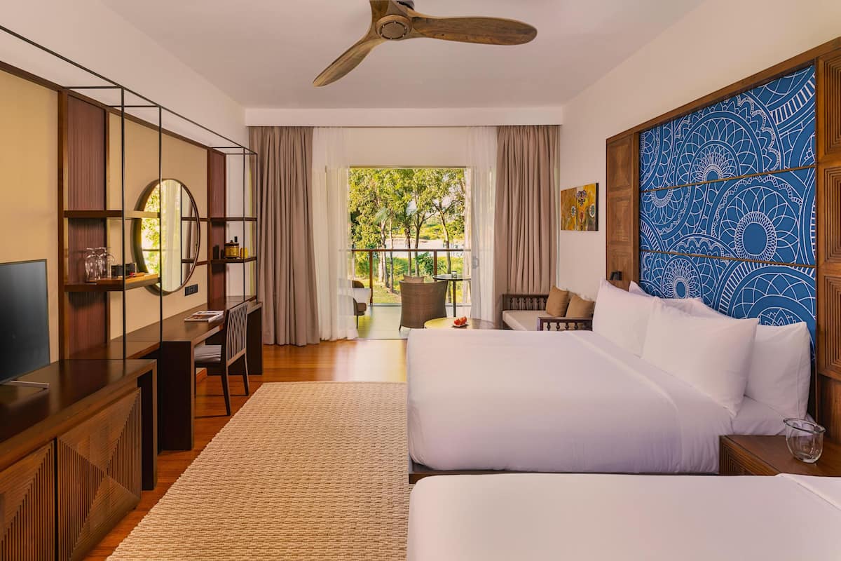 A hotel room with a large bed, a desk with a TV, a balcony with a view of greenery, and a ceiling fan.