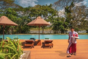 A poolside area with lounge chairs and umbrellas, a person carrying a tray with drinks, and a view of a natural landscape.