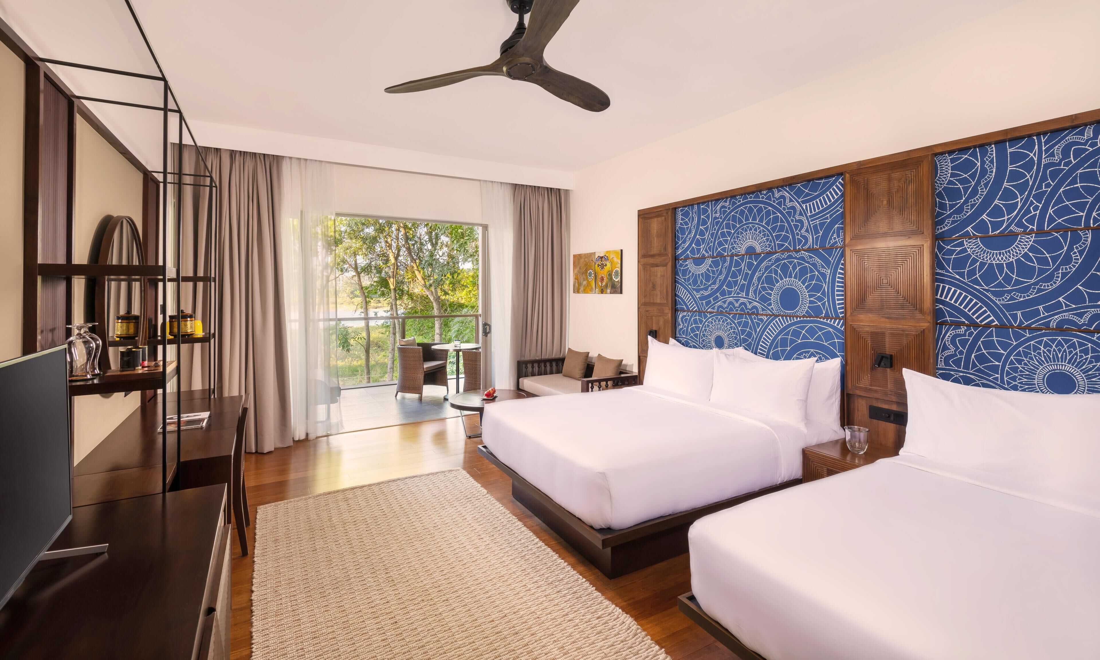 A hotel room with two beds, a ceiling fan, a TV, a desk, and a view of greenery.