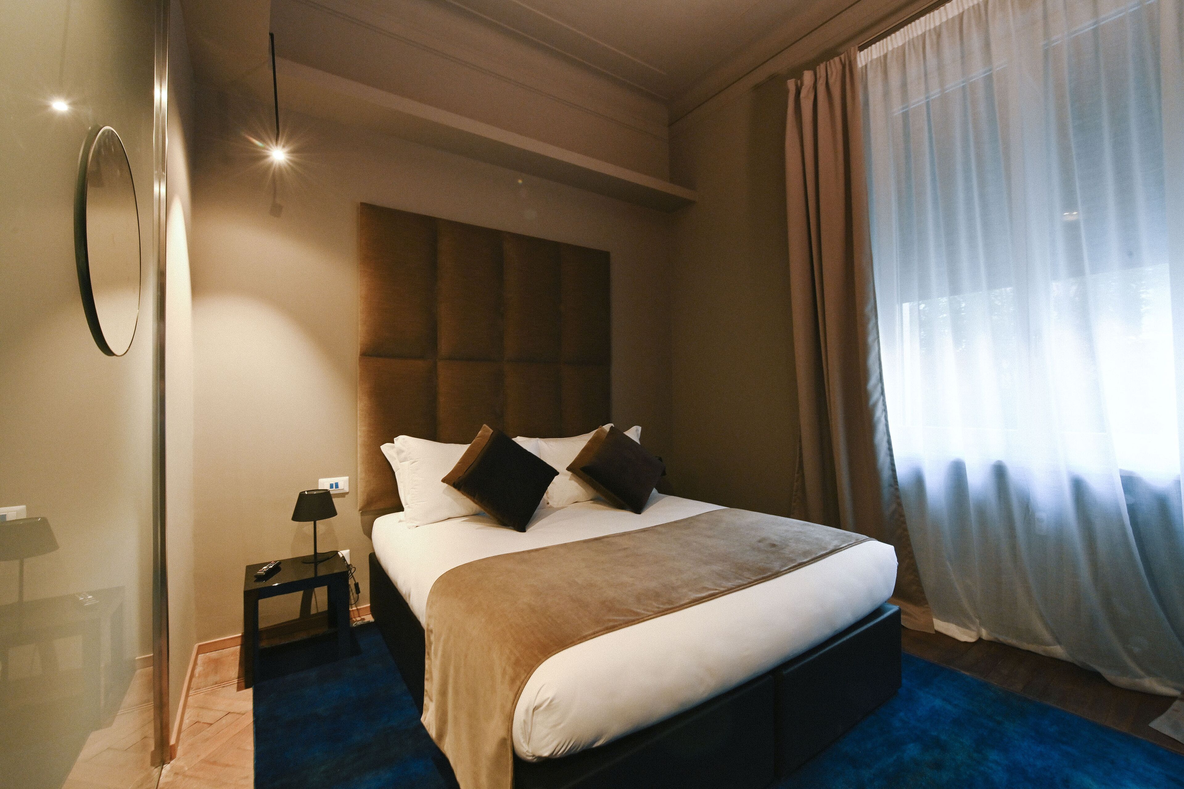 A hotel room with a large bed, a bedside table with a lamp, a mirror on the wall, and a window with sheer curtains.