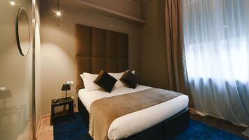 A hotel room with a large bed, a bedside table with a lamp, a mirror on the wall, and a window with sheer curtains.