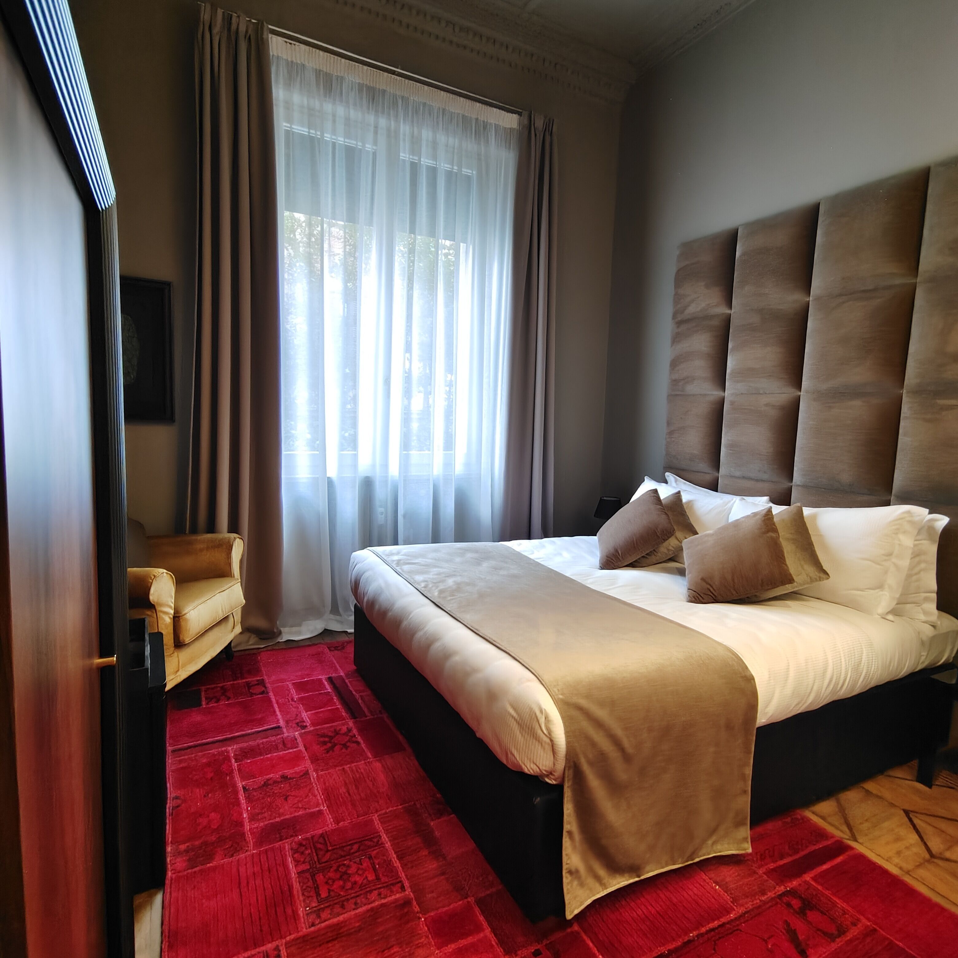 A hotel room with a large bed, a red patterned carpet, a sofa, and a window with sheer curtains.