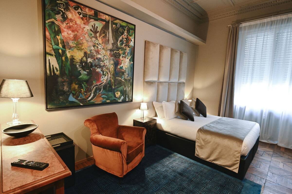 A hotel room with a large abstract painting, a bed with a beige cover, a brown armchair, a nightstand with a lamp, and a remote control on a table.