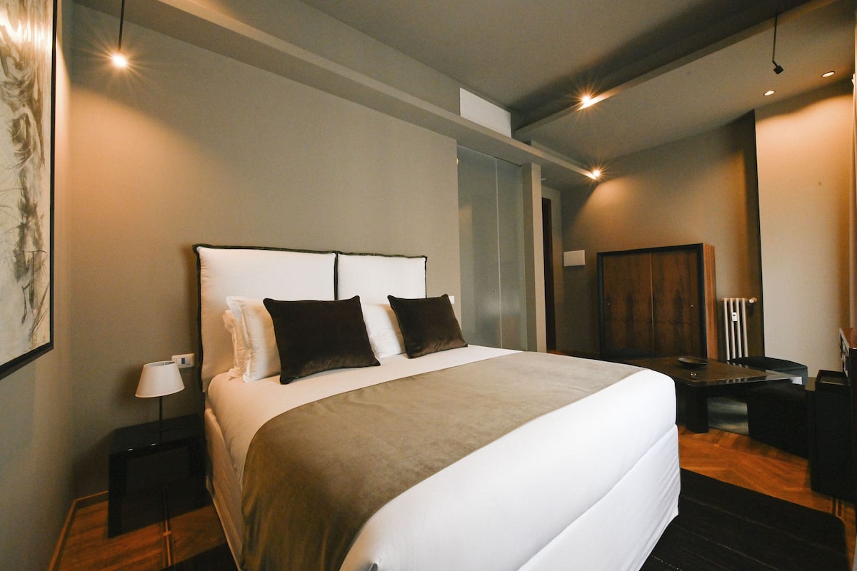 A modern hotel room with a large bed, bedside table, and a desk with a chair.