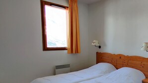 A simple, clean room with a bed, a window with curtains, a wall-mounted lamp, and a radiator. - 3-room apartment, balcony, pets allowed (Le Dévoluy)