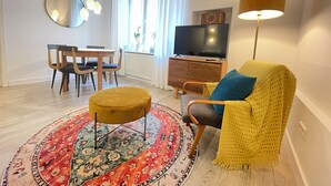 En moderne stue med et rundt tæppe, en gul fodskammel, en sofa med et gult pled, et fjernsyn på et træskab, samt et spiseområde med stole og bord.