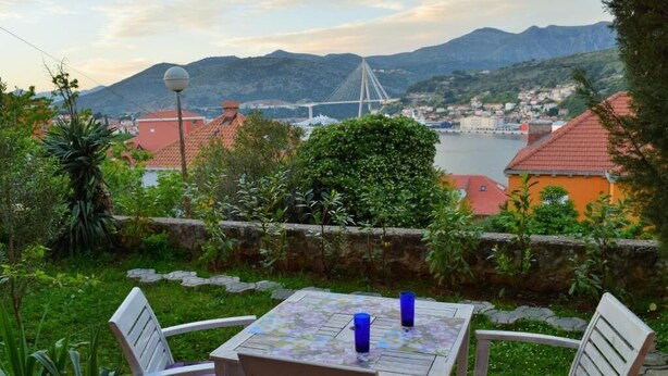 A table with two chairs and two blue glasses, set against a backdrop of a mountainous landscape and a bridge. - Leela Apartment (Dubrovnik)