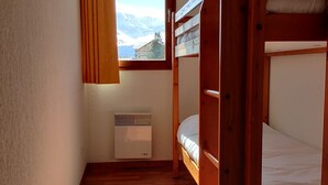 Une petite pièce chaleureuse avec des lits superposés en bois, une fenêtre avec des rideaux et un radiateur fixé au mur.