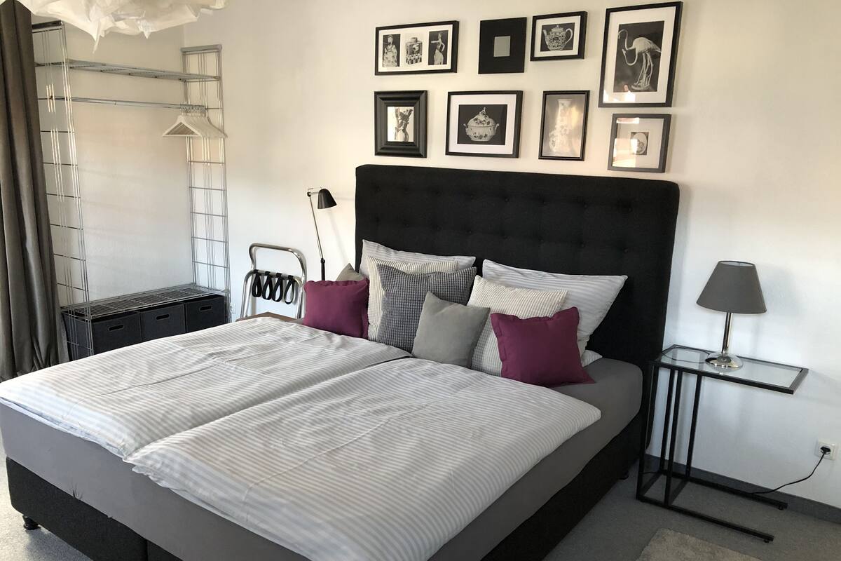 A neatly made bed with striped bedding, a dark headboard, and several pillows. There is a small bedside table with a lamp, and a wall with framed pictures.