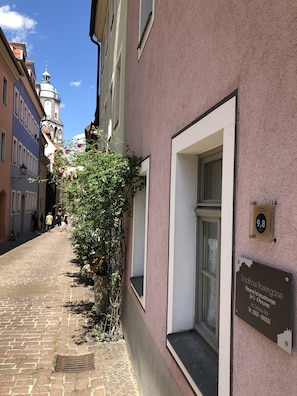A narrow cobblestone street with a building featuring a plaque and a window with a view.