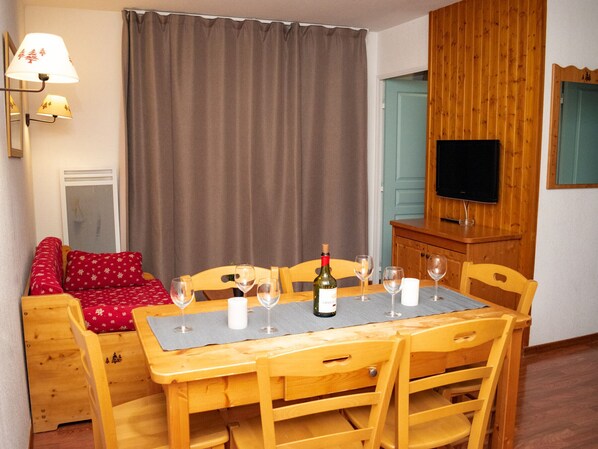 A cozy room with a wooden dining table set for four, a sofa with red pillows, a television on a wooden cabinet, and a large window with curtains. - 2-room apartment in Superdevoluy for 4 people with balcony (Le Dévoluy)